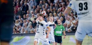 Ein Handballspieler in einem weißen Trikot bereitet sich darauf vor, den Ball während eines Spiels zu fangen. Zu sehen sind Mannschaftskameraden, ein Gegner und ein Schiedsrichter; im Hintergrund sieht eine Zuschauermenge zu.