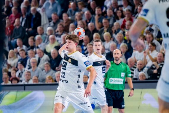 Ein Handballspieler in einem weißen Trikot bereitet sich darauf vor, den Ball während eines Spiels zu fangen. Zu sehen sind Mannschaftskameraden, ein Gegner und ein Schiedsrichter; im Hintergrund sieht eine Zuschauermenge zu.