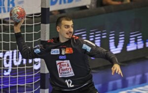 Ein Handballtorwart in einem schwarzen Trikot mit der Nummer 12 macht sich bereit, den Ball zu werfen. Er steht vor dem Tor, blickt nach links, hebt den Arm und hält den Ball fest.
