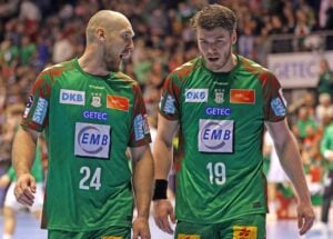 Zwei männliche Handballspieler in grünen Trikots mit den Nummern 24 und 19 laufen nebeneinander auf dem Spielfeld und scheinen sich zu unterhalten. Der Hintergrund zeigt eine unscharfe Menschenmenge und andere Spieler.