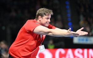 Ein Mann in einem roten Hemd mit einer Trillerpfeife und einem Ausweis streckt beide Arme nach vorne aus, die Handflächen nach oben, als ob er während einer Sportveranstaltung ein Signal geben oder Anweisungen erteilen würde. Der Hintergrund ist unscharf, Zuschauer und Lichter sind zu sehen.