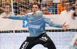 Ein Handballtorwart in einem blauen Trikot und einer schwarzen Hose steht mit ausgebreiteten Armen, konzentriert auf einen herannahenden Ball während eines Spiels, mit einem Netz und Zuschauern im Hintergrund.