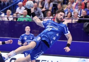 Ein männlicher Handballspieler in einem blauen Trikot springt mit ausgestrecktem Arm in die Luft, um den Ball zu werfen. Die Zuschauer schauen von der Tribüne im Hintergrund zu.