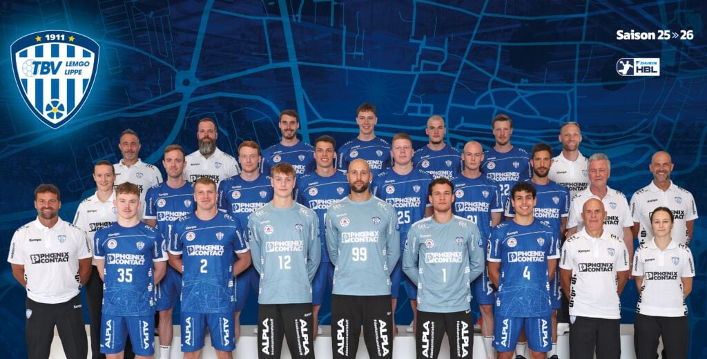 25_vor_TBV_Teambild Eine Herren-Handballmannschaft posiert in blauen und weißen Trikots mit Trainern und Mitarbeitern, die auf beiden Seiten stehen. Im Hintergrund sind eine blaue Landkarte und das Mannschaftslogo zu sehen. Der Text