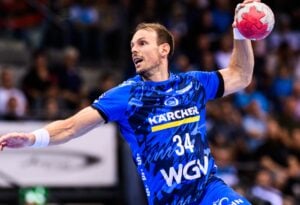 Ein männlicher Handballspieler in einem blauen Trikot mit der Nummer 34 bereitet sich während eines Spiels auf den Wurf vor, während die Zuschauer im Hintergrund verschwommen zu sehen sind.