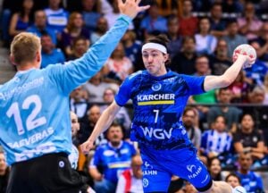 Ein männlicher Handballspieler in einem blauen Trikot bereitet sich darauf vor, den Ball zu werfen, während ein Torwart in einem hellblauen Trikot seinen Arm zum Blocken hebt.