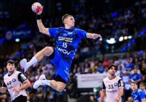 Ein männlicher Handballspieler in einem blauen Trikot wirft den Ball in der Luft, während Spieler in weißen und schwarzen Trikots in einer überfüllten Halle zusehen.