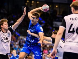 Ein männlicher Handballspieler in einem blauen Trikot springt, um den Ball zu werfen, während er von drei Spielern in weißen Trikots während eines Spiels in einer überfüllten Halle blockiert wird.