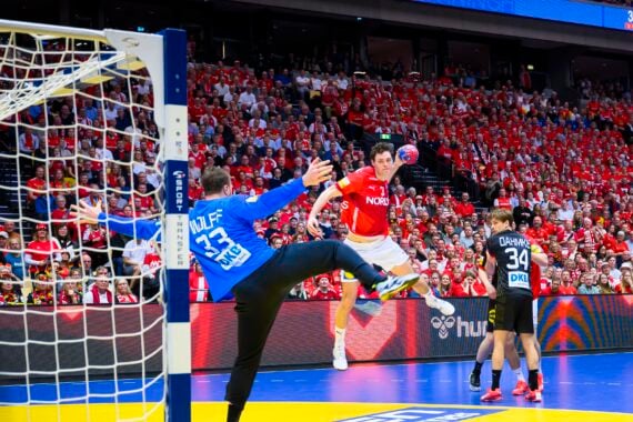 Ein Handballspieler in einem rot-weißen Trikot springt zum Schuss, während ein Torwart in Blau versucht, den Schuss zu blocken; eine vollbesetzte Arena mit Fans in Rot schaut gebannt zu.