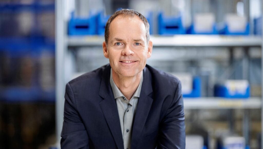 26_Kai_Kruse_Titel Ein Mann in einem dunklen Blazer und einem hellen Hemd lächelt, während er in einem gut beleuchteten Innenraum vor Metallregalen mit blauen Lagerbehältern sitzt.