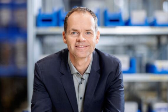 Ein Mann in einem dunklen Blazer und einem hellen Hemd lächelt, während er in einem gut beleuchteten Innenraum vor Metallregalen mit blauen Lagerbehältern sitzt.