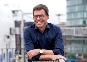 Ein Mann mit Brille und dunkelblauem Hemd lächelt, während er sich an ein Geländer im Freien lehnt, mit unscharfen modernen Gebäuden im Hintergrund.