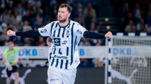 Ein männlicher Handballspieler in einem weiß-schwarz gestreiften Trikot mit ausgestreckten Armen feiert während eines Spiels, mit einem unscharfen Schiedsrichter und Publikum im Hintergrund.