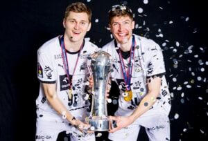 Zwei männliche Athleten in weißen Uniformen lächeln und halten eine große silberne Trophäe zusammen, während sie Medaillen um den Hals tragen und vor einem dunklen Hintergrund Konfetti fällt.