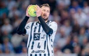Ein männlicher Handballspieler in einem schwarz-weißen Trikot bereitet sich während eines Spiels auf den Wurf eines gelben Balls vor, mit einer unscharfen Menschenmenge im Hintergrund.