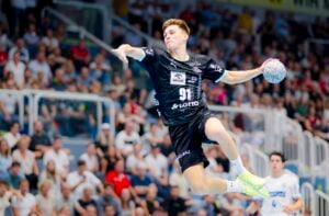 Ein männlicher Handballspieler in einem schwarzen Trikot springt in der Luft, um den Ball während eines Spiels zu werfen, während im Hintergrund eine unscharfe Menge von der Tribüne aus zusieht.