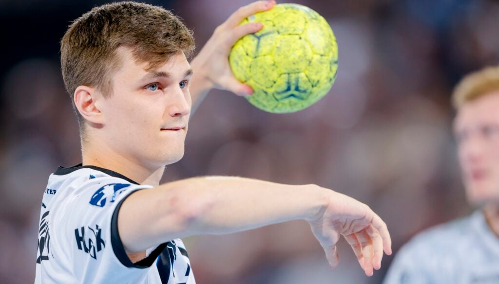HANDBALL 2025/26: THW Kiel - Frisch Auf! Göppingen Ein junger männlicher Handballspieler in einem weißen Trikot bereitet sich auf den Wurf eines gelben Handballs vor. Er ist konzentriert, hat den linken Arm ausgestreckt und hält den Ball mit dem rechten Arm. Der Hintergrund zeigt eine unscharfe Menge und einen anderen Spieler.