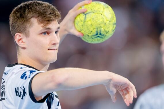 Ein junger männlicher Handballspieler in einem weißen Trikot bereitet sich auf den Wurf eines gelben Handballs vor. Er ist konzentriert, hat den linken Arm ausgestreckt und hält den Ball mit dem rechten Arm. Der Hintergrund zeigt eine unscharfe Menge und einen anderen Spieler.