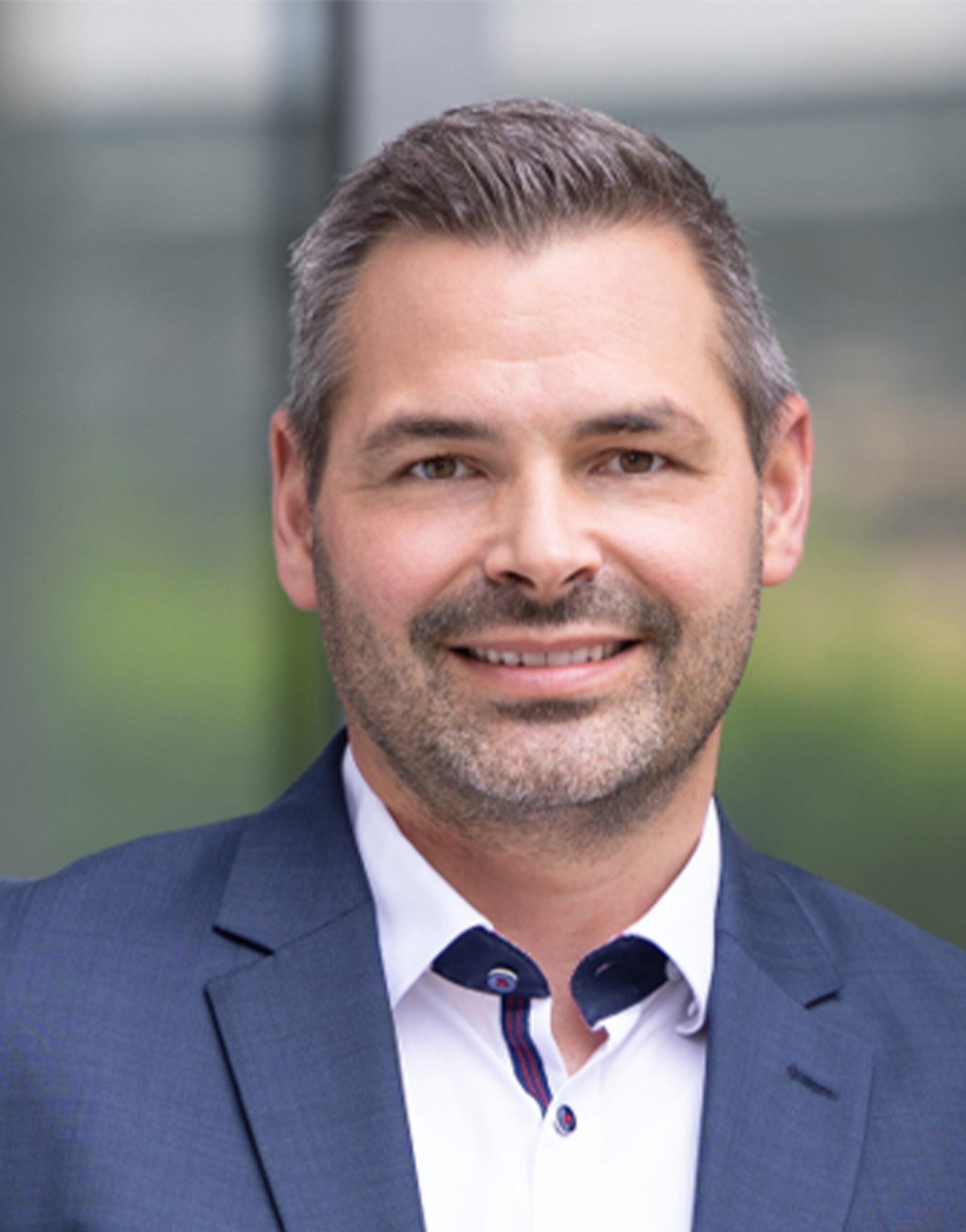 AR_Dennis_Kuhlbach Ein lächelnder Mann mit kurzem dunklem Haar und gestutztem Bart, der eine blaue Anzugsjacke über einem weißen Hemd trägt, steht vor einem unscharfen Hintergrund im Freien.