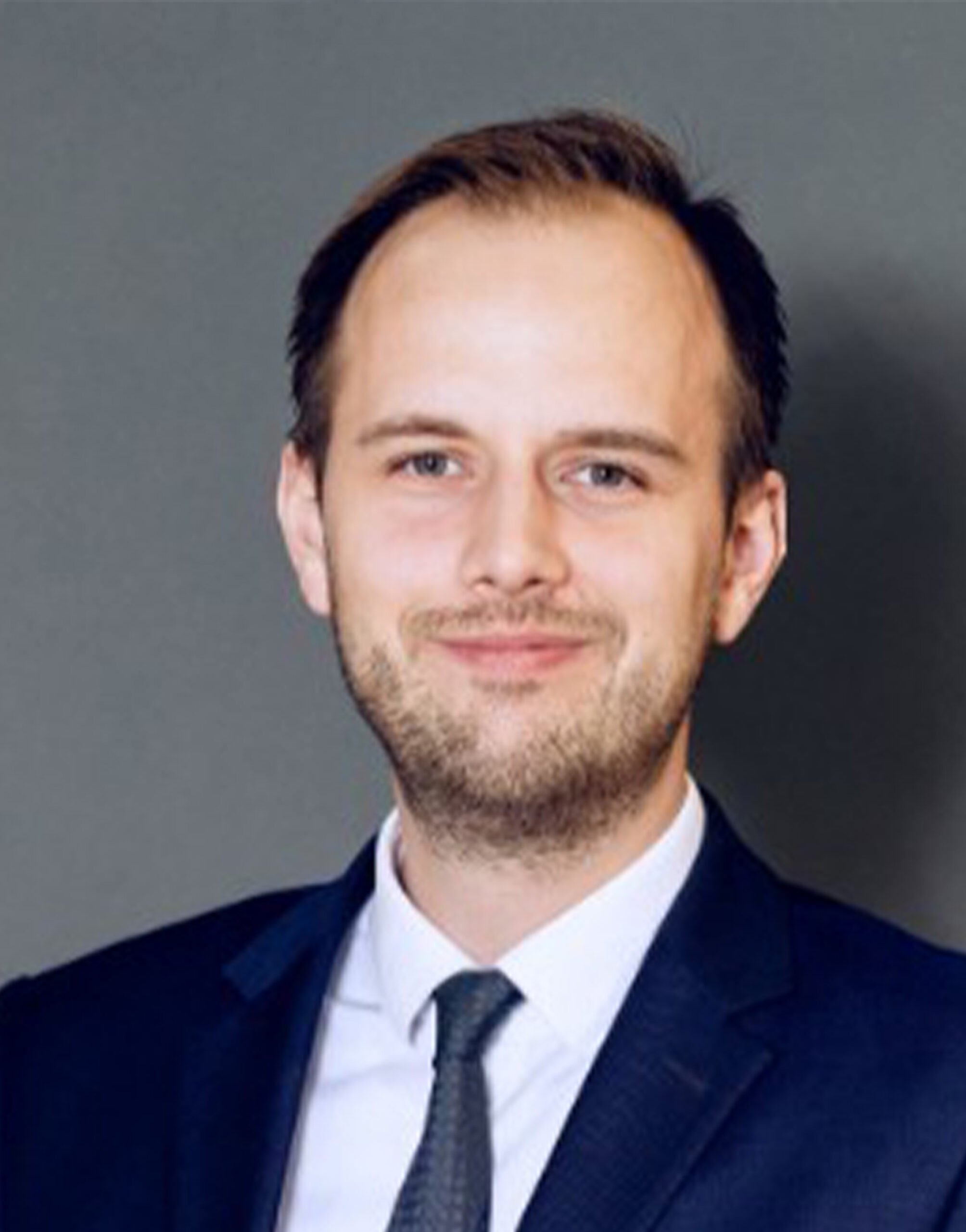 AR_Jan-Phlipp_Petersen Ein Mann in einem dunklen Anzug, weißem Hemd und Krawatte lächelt sanft in die Kamera. Er hat kurzes braunes Haar und einen gestutzten Bart. Hinter ihm ist ein neutraler grauer Hintergrund zu sehen.