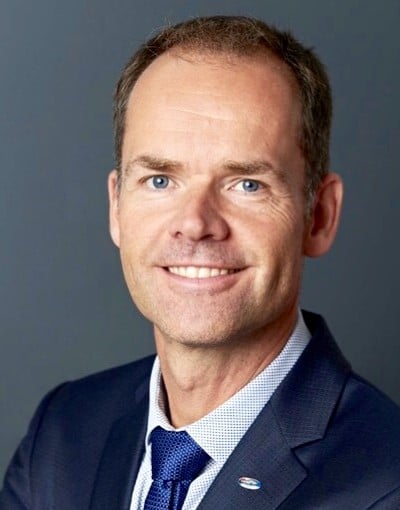 AR_Kai_Kruse Ein Mann in einem dunklen Anzug, weißem Hemd und blauer Krawatte lächelt in die Kamera vor einem schlichten grauen Hintergrund. Er hat kurzes braunes Haar und blaue Augen.