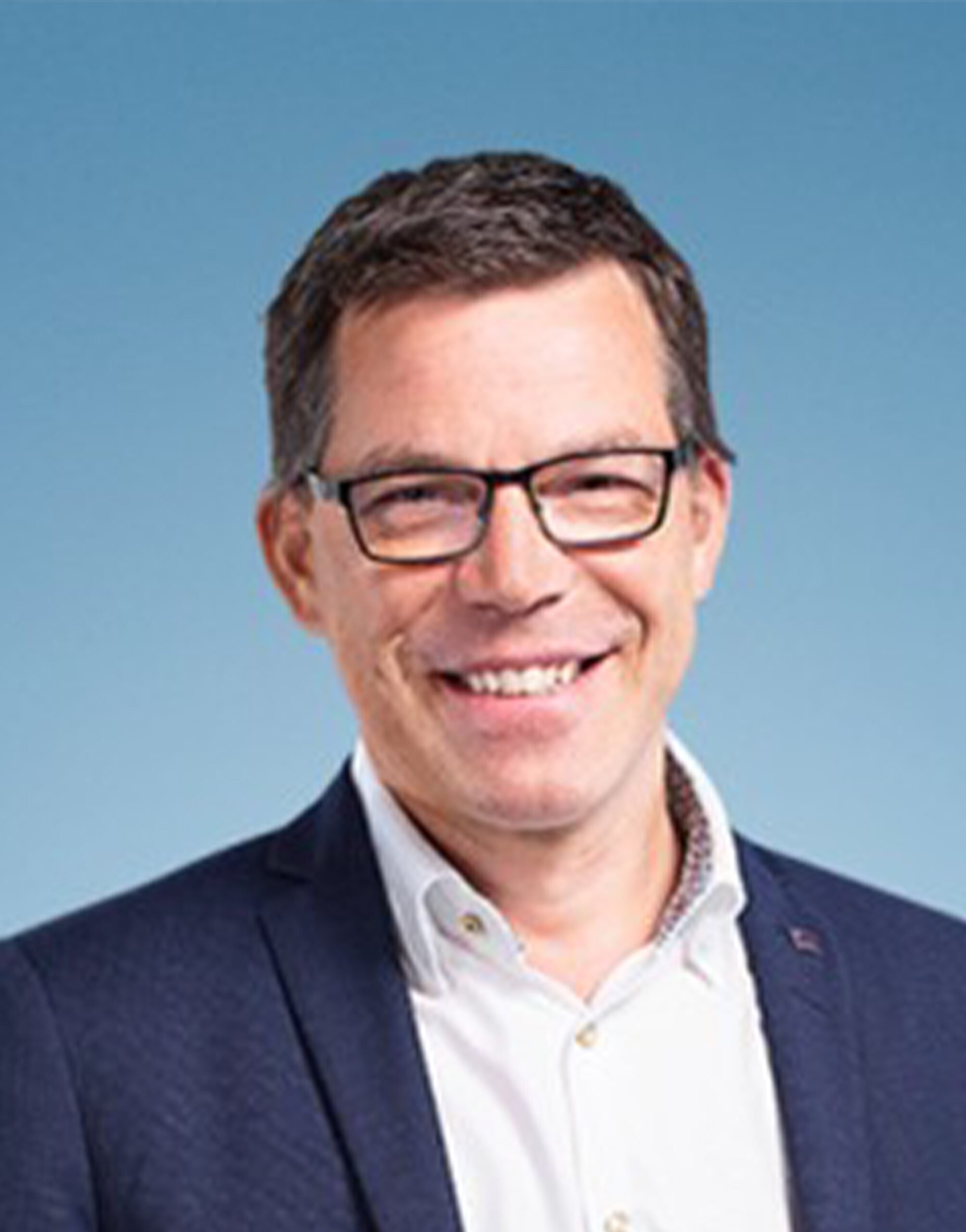 AR_Knud_Hansen Ein Mann mit kurzen braunen Haaren und Brille, der eine dunkle Anzugsjacke über einem weißen Kragenhemd trägt, lächelt vor einem hellblauen Hintergrund.