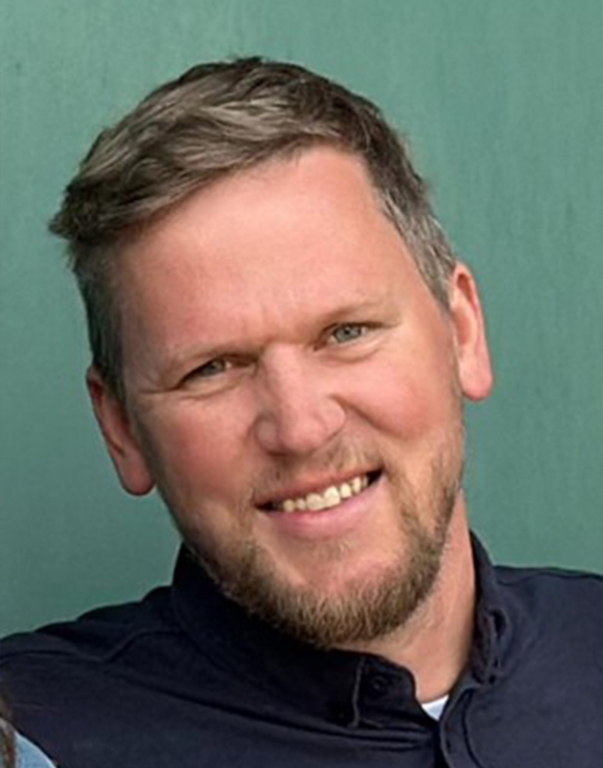 AR_Lasse_Elwardt Ein Mann mit kurzen hellbraunen Haaren und einem Bart, der ein dunkles Hemd mit Kragen trägt, lächelt vor einem einfarbig grünen Hintergrund.