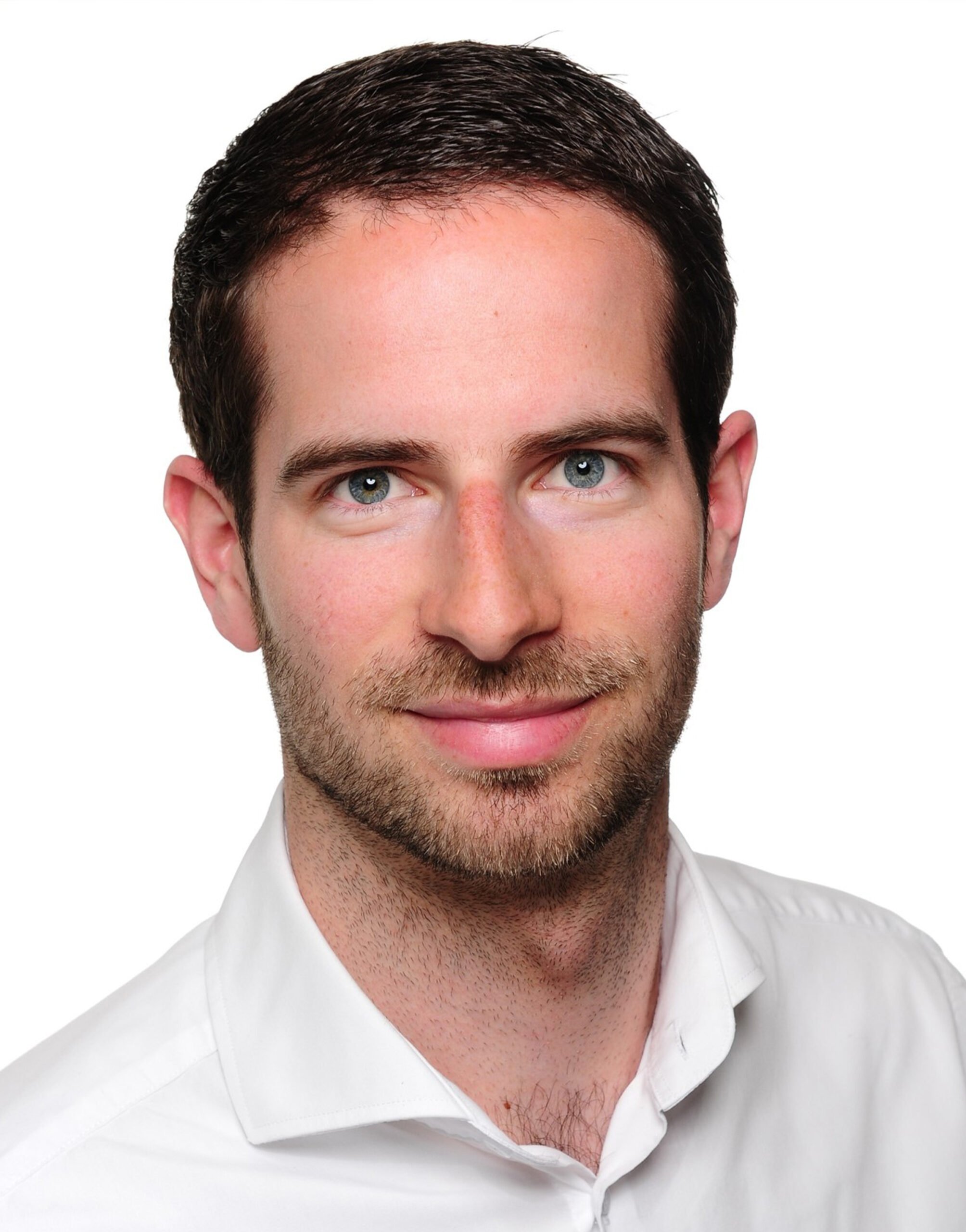 AR_Ole_Bischoff Ein Mann mit kurzem braunem Haar, gestutztem Bart und blauen Augen, der ein weißes Hemd mit Kragen trägt, lächelt in die Kamera vor einem schlichten weißen Hintergrund.