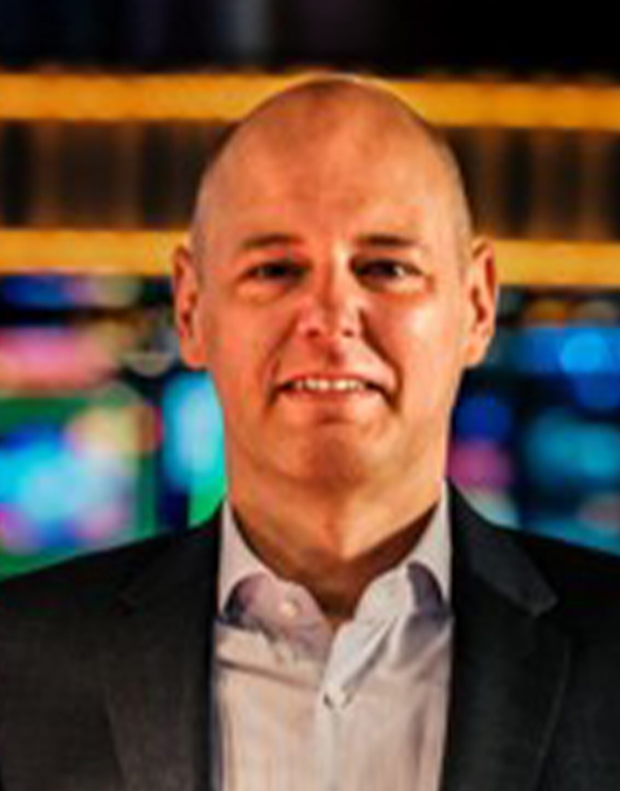 AR_Rainer_Schepull Ein Mann mittleren Alters mit Glatze, Anzugjacke und Kragenhemd, steht vor einem bunten, verschwommenen Hintergrund aus Lichtern oder Bildschirmen.