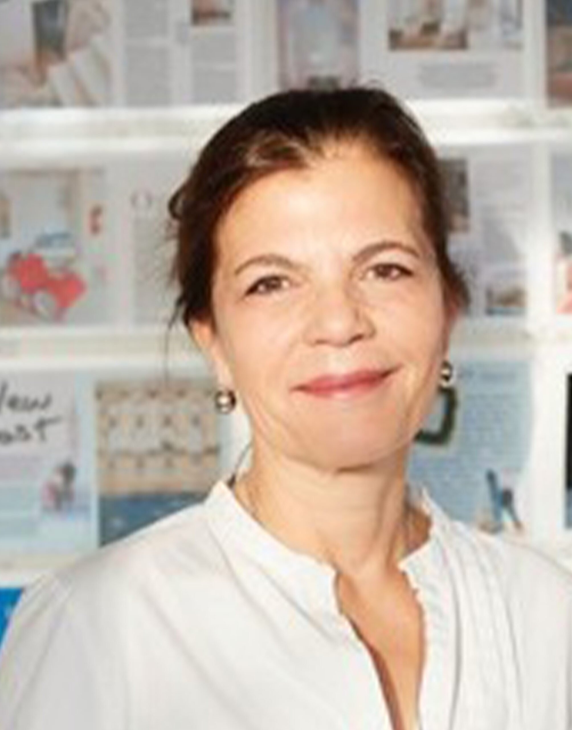 AR_Susan_Molzow Eine Frau mit zurückgebundenem braunem Haar, weißer Bluse und kleinen Ohrringen steht vor einer mit Zeitschriftenseiten und Fotos bedeckten Wand und lächelt sanft in die Kamera.