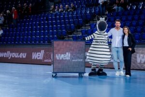 Zwei Personen und ein Zebra-Maskottchen stehen zusammen auf einem Indoor-Basketballplatz in der Nähe einer digitalen Anzeigetafel. Im Hintergrund sind leere blaue Sitze und ein paar Zuschauer zu sehen.