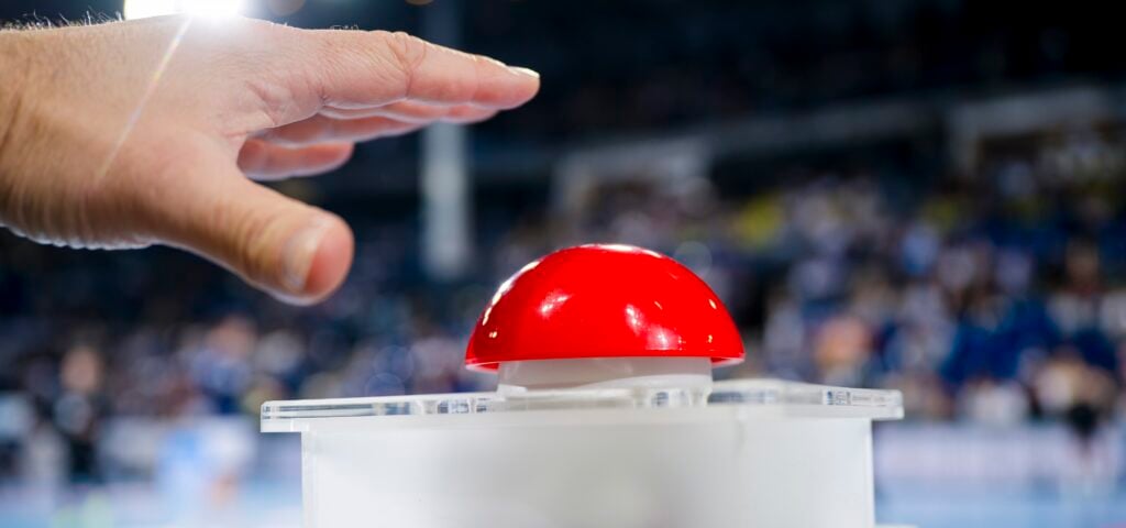 Eine Hand schwebt über einem großen roten Knopf auf weißem Grund, mit einer unscharfen Menschenmenge im Hintergrund.