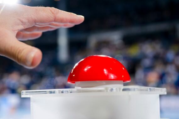 Eine Hand schwebt über einem großen roten Knopf auf weißem Grund, mit einer unscharfen Menschenmenge im Hintergrund.