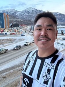 Ein Mann in einem schwarz-weiß gestreiften Sporttrikot lächelt in die Kamera, während im Hintergrund eine verschneite Stadt, Straßen, Autos und Berge zu sehen sind.