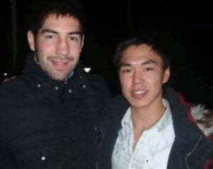 Zwei junge Männer stehen bei Nacht dicht beieinander und lächeln in die Kamera. Einer trägt eine dunkle Jacke, der andere ein helles Hemd unter einem dunklen Mantel mit einer pelzgefütterten Kapuze. Der Hintergrund ist dunkel und nicht klar erkennbar.