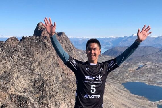 Eine lächelnde Person steht mit erhobenen Armen auf einem felsigen Berggipfel und trägt ein schwarzes Sporttrikot. Dahinter sind schroffe Berge, blauer Himmel und Seen im darunter liegenden Tal zu sehen.