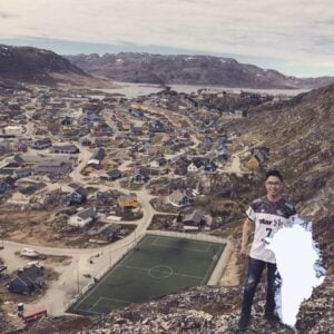 Eine Person steht auf einem felsigen Hügel und überblickt eine kleine Stadt mit bunten Häusern, Bergen und einem Fußballfeld. Ein Umriss von Grönland erscheint in der unteren rechten Ecke.