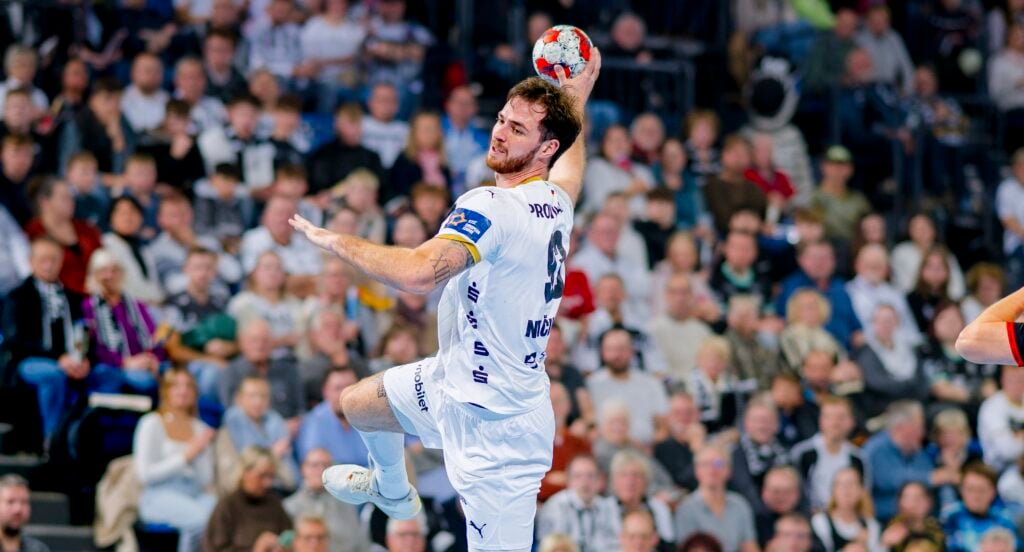 HANDBALL 2025/26: THW Kiel - BSV Bern Ein männlicher Handballspieler in einem weißen Trikot springt in der Luft, bereit, den Ball zu werfen. Im Hintergrund ist eine große, verschwommene Menschenmenge zu sehen, die das Geschehen in einer Sportarena verfolgt.
