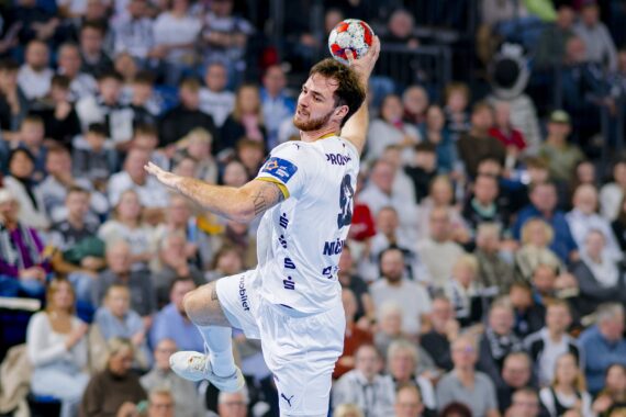 Ein männlicher Handballspieler in einem weißen Trikot springt in der Luft, bereit, den Ball zu werfen. Im Hintergrund ist eine große, verschwommene Menschenmenge zu sehen, die das Geschehen in einer Sportarena verfolgt.