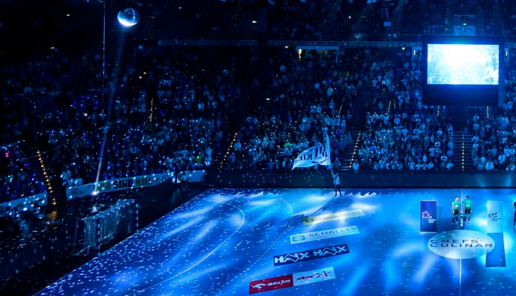 Eine große, mit Zuschauern gefüllte Sporthalle, beleuchtet von blauen Lichtern. Sportlerinnen und Sportler stehen mit einer weißen Fahne auf dem Spielfeld, ein Scheinwerfer strahlt und Konfetti fliegt - eine elektrisierende Szene bei der THW Kiel Handball-Bundesliga Saison 2025/2026.