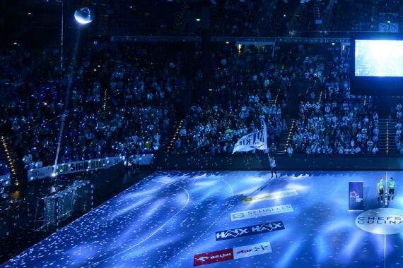 Eine große, mit Zuschauern gefüllte Sporthalle, beleuchtet von blauen Lichtern. Sportlerinnen und Sportler stehen mit einer weißen Fahne auf dem Spielfeld, ein Scheinwerfer strahlt und Konfetti fliegt - eine elektrisierende Szene bei der THW Kiel Handball-Bundesliga Saison 2025/2026.