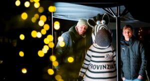 Zwei Männer in Winterjacken stehen neben einer Person in einem Zebra-Maskottchen-Kostüm unter einem nächtlichen Vordach, wobei gelbe Bokeh-Lichter im Vordergrund verschwimmen.