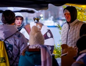 Eine Gruppe von Menschen in Winterkleidung versammelt sich um ein sich drehendes Preisrad an einem Marktstand im Freien. Lichter und ein geschmückter Baum schaffen eine festliche Atmosphäre. Ein Mann in einer Kapuzenweste steht neben dem Rad und beaufsichtigt die Aktivität.