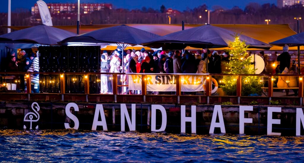 Eine Gruppe von Menschen versammelt sich am Abend an einem festlichen Ort am Flussufer namens SANDHAFEN, wo warme Lichter, große Sonnenschirme, ein beleuchteter Weihnachtsbaum und ein Wir sind Kiel-Banner zu sehen sind.