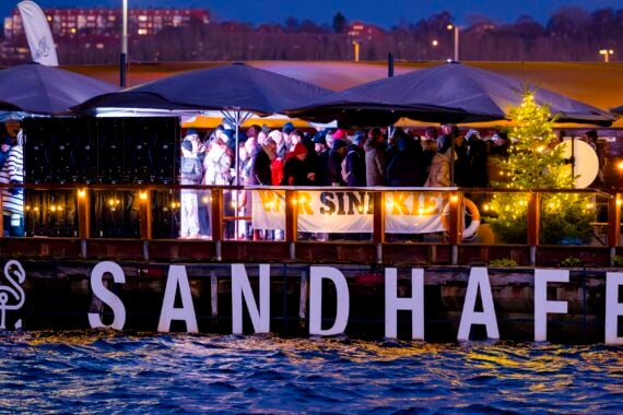 Eine Gruppe von Menschen versammelt sich am Abend an einem festlichen Ort am Flussufer namens SANDHAFEN, wo warme Lichter, große Sonnenschirme, ein beleuchteter Weihnachtsbaum und ein Wir sind Kiel-Banner zu sehen sind.