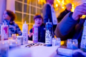 Kinder sitzen an einem hell erleuchteten Tisch mit Flaschen mit bunten Farben und Kunstzubehör und beschäftigen sich mit einer kreativen Tätigkeit. Die Szene ist warm und festlich, und im Hintergrund hängen goldene Lichter.