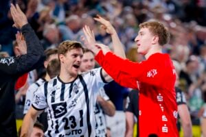 Zwei männliche Handballspieler, einer in einem schwarz-weiß gestreiften Trikot, der andere in einem roten Trikot, feiern gemeinsam mit High-Fives, lächelnd und umgeben von jubelnden Mannschaftskameraden und einer verschwommenen Menge im Hintergrund.