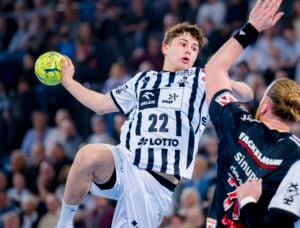 Ein männlicher Handballspieler in einem weiß-schwarz gestreiften Trikot springt, um einen gelben Ball zu werfen, während ein Verteidiger in einem schwarzen Trikot versucht, ihn zu blockieren. Im Hintergrund sieht man eine unscharfe Zuschauermenge, die das Spiel verfolgt.