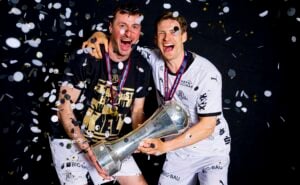 Zwei lächelnde Athleten in Sportuniformen feiern, halten eine große silberne Trophäe und tragen Medaillen, während um sie herum vor einem schwarzen Hintergrund Konfetti fällt.