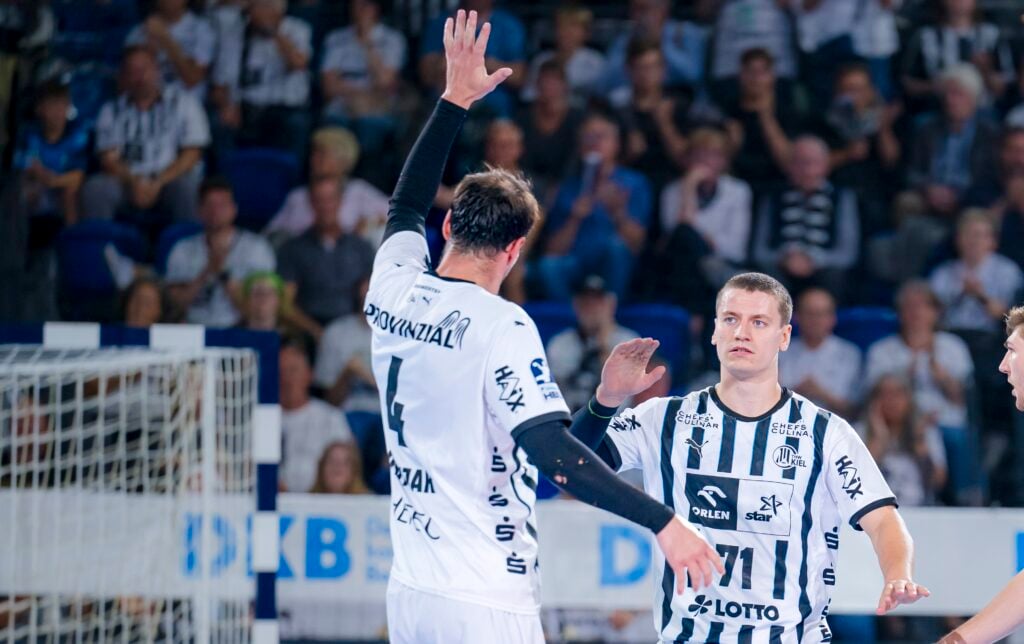 Zwei männliche Handballspieler in schwarz-weiß gestreiften Trikots stehen auf einem Hallenplatz. Ein Spieler hebt die Hand zum Abklatschen, während der andere ihn anschaut. Im unscharfen Hintergrund sind Zuschauer zu sehen.